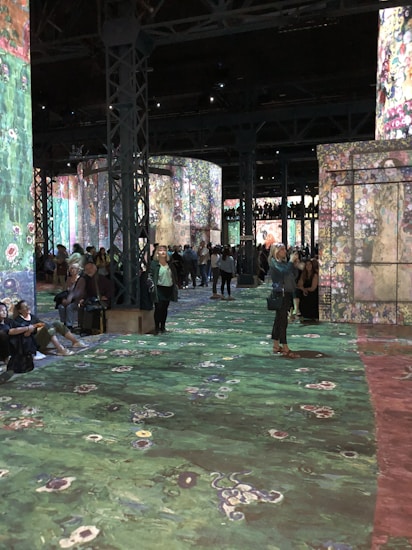 A large indoor exhibition space with projections of colorful floral and abstract patterns on walls and floors. The room is filled with people, some sitting and others walking, engaging with the immersive art experience. Tall metal columns are present, supporting the structure.