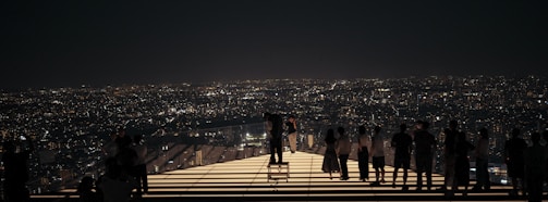 Groups of people stand on an illuminated platform overlooking a sprawling cityscape at night, with countless lights twinkling across the horizon.