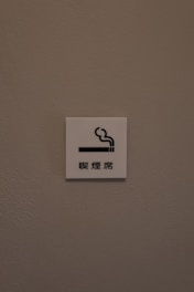 A small white sign is mounted on a textured, light brown wall. The sign features a simple graphic of a cigarette with smoke rising from it, accompanied by Chinese characters underneath.