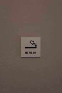 A small white sign is mounted on a textured, light brown wall. The sign features a simple graphic of a cigarette with smoke rising from it, accompanied by Chinese characters underneath.
