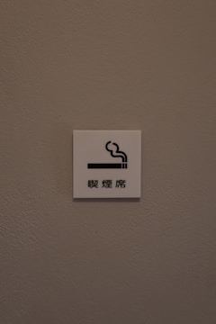 A small white sign is mounted on a textured, light brown wall. The sign features a simple graphic of a cigarette with smoke rising from it, accompanied by Chinese characters underneath.