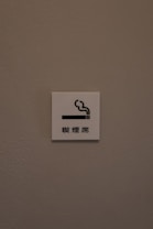 A small white sign is mounted on a textured, light brown wall. The sign features a simple graphic of a cigarette with smoke rising from it, accompanied by Chinese characters underneath.