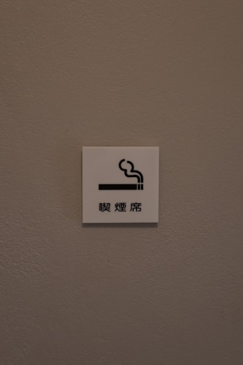 A small white sign is mounted on a textured, light brown wall. The sign features a simple graphic of a cigarette with smoke rising from it, accompanied by Chinese characters underneath.