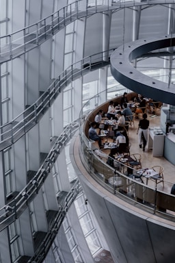 Curved architectural design featuring multiple levels with large windows, a modern interior characterized by steel and glass elements. A circular dining area with people seated at tables, engaged in conversation or dining activities.