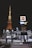 A brightly lit tower rises against the night sky, with a gas station in the foreground displaying signs in Japanese. The gas station is marked by the ENEOS logo, and there are a couple of people walking nearby. The lit tower provides a warm contrast to the darker surroundings.