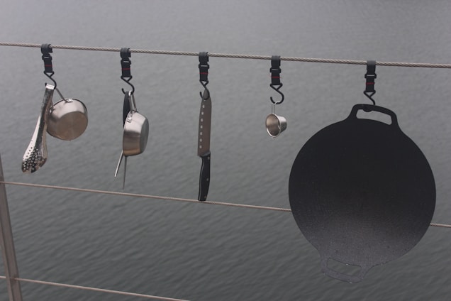 Various kitchen utensils hang from a cable above a body of water. There is a ladle, a saucepan, a cheese grater, a kitchen knife, and a cast iron griddle, all attached to the cable with individual hooks.