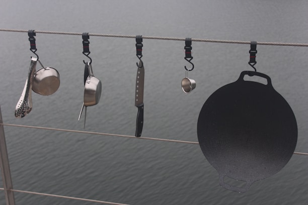 Various kitchen utensils hang from a cable above a body of water. There is a ladle, a saucepan, a cheese grater, a kitchen knife, and a cast iron griddle, all attached to the cable with individual hooks.