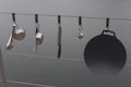 Various kitchen utensils hang from a cable above a body of water. There is a ladle, a saucepan, a cheese grater, a kitchen knife, and a cast iron griddle, all attached to the cable with individual hooks.
