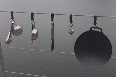 Various kitchen utensils hang from a cable above a body of water. There is a ladle, a saucepan, a cheese grater, a kitchen knife, and a cast iron griddle, all attached to the cable with individual hooks.
