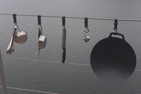 Various kitchen utensils hang from a cable above a body of water. There is a ladle, a saucepan, a cheese grater, a kitchen knife, and a cast iron griddle, all attached to the cable with individual hooks.