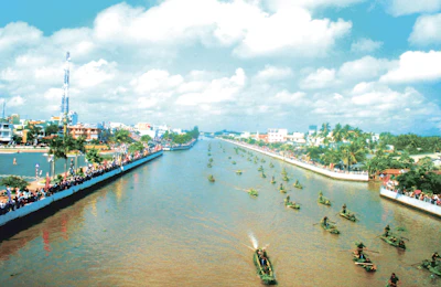 Cheering crowd on the riverbank waving flags as boats race by in vibrant colors.