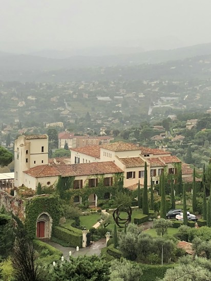 A large, elegant villa with beige walls and red-tiled roofs is surrounded by lush greenery and tall cypress trees. The landscape features manicured gardens and a circular sculpture on the lawn. Positioned against a backdrop of rolling hills and a hazy sky, the villa overlooks a distant town nestled in a valley.
