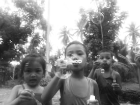 Children are outdoors blowing soap bubbles, with trees and foliage in the background. Three kids are visible in focus, each holding a bubble wand and looking at the bubbles they’ve created.