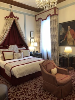 A luxurious bedroom with a regal design features a large bed with ornate red and white bedding. A decorative canopy is suspended above the bed, adorned with matching curtains. The room is elegantly furnished with a plush armchair and detailed carpet with intricate patterns. A chandelier hangs from the ceiling, adding to the opulence. One wall is decorated with a large portrait of a person dressed in historical attire. Soft, ambient light emanates from lamps placed on bedside tables.