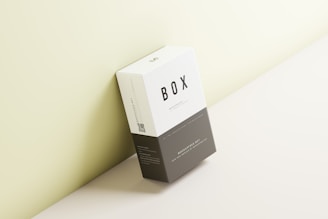 A rectangular box is placed against a wall with a minimalist design. The box has a clean, modern look with a white top half and a dark gray bottom half. The word 'BOX' is prominently printed on the front. The background is a soft light green hue.