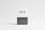 A minimalistic box with a clean white and gray design. The box is labeled with the word 'BOX' in bold black letters on the upper portion, and more text in smaller font is seen below. The background is light gray, creating a simple and modern aesthetic.
