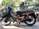 A black motorbike with a Honda logo and a basket attached to the front. The bike is parked on a textured surface surrounded by plants in the background, with a focus on the detailed design of the seat and red suspension coil.