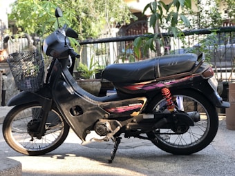 A black motorbike with a Honda logo and a basket attached to the front. The bike is parked on a textured surface surrounded by plants in the background, with a focus on the detailed design of the seat and red suspension coil.