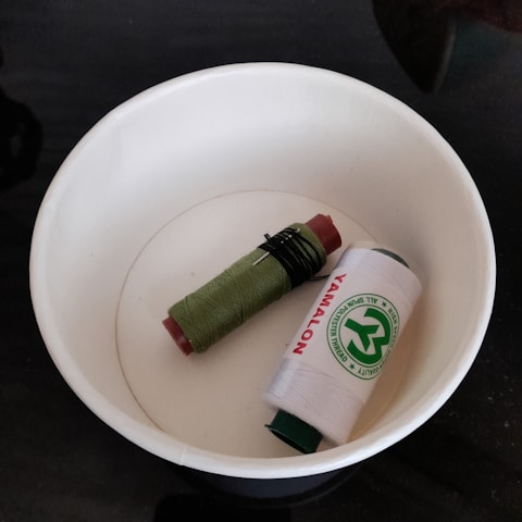 A white plastic bowl containing two spools of thread, one green and one white. The white spool has a green label with text and a logo on it.