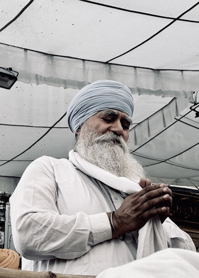An elderly man with a long beard and mustache is wearing a turban and traditional clothing. He has his eyes closed and hands together, possibly in prayer or reflection. The background shows a partially transparent canopy with a geometric pattern.