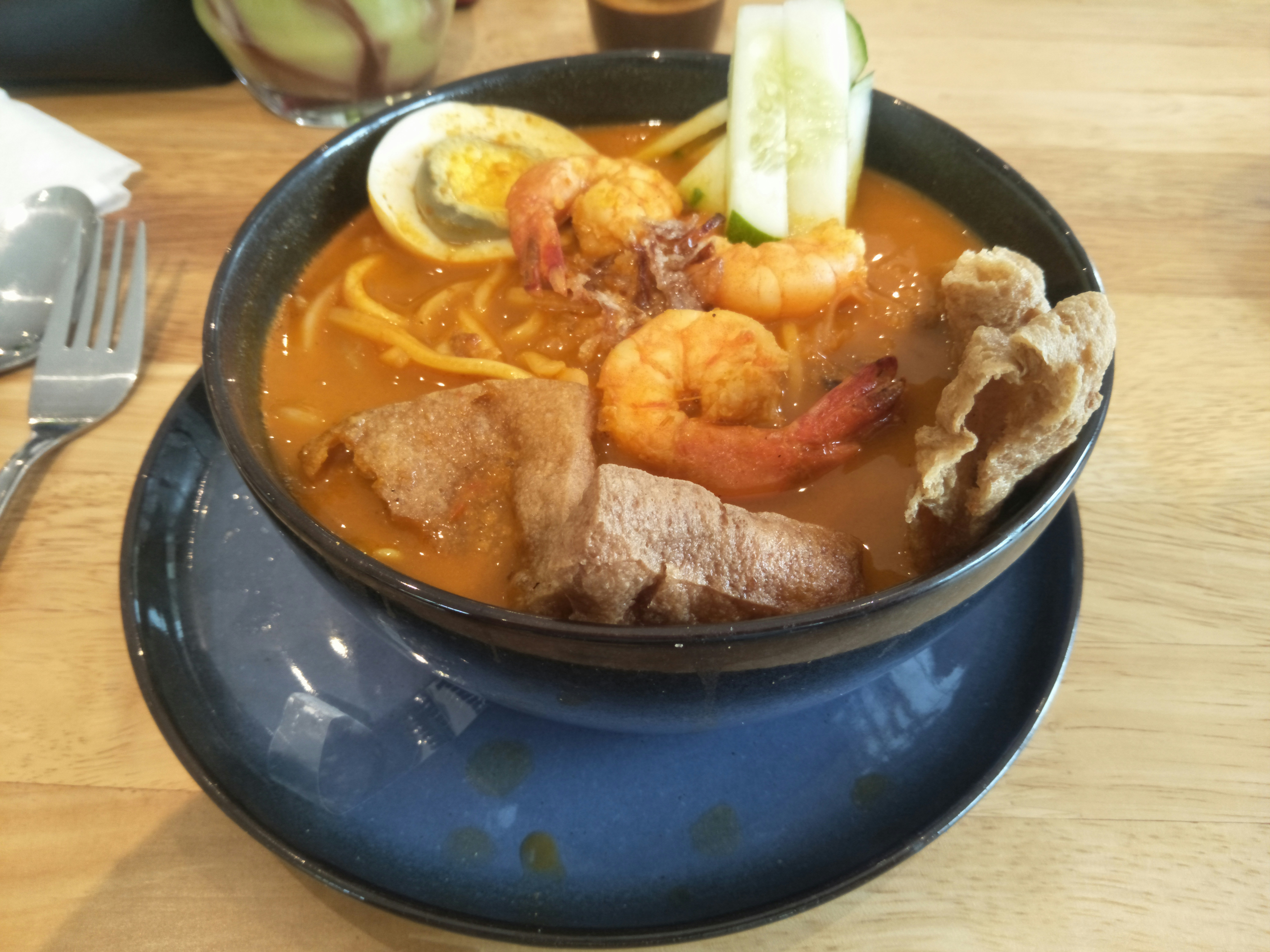 a bowl of yellow noodles served with a spicy sauce and topped with meat egg shrimp and cucumber. these noodles are also served with a mixture of tofu and onion granules along with quality spices.