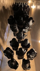 A collection of black sculptures resembling gorillas is displayed in a vertical, linear arrangement inside a narrow corridor. The sculptures are suspended from the ceiling, casting shadows due to the warm lighting from the lamps on the walls.