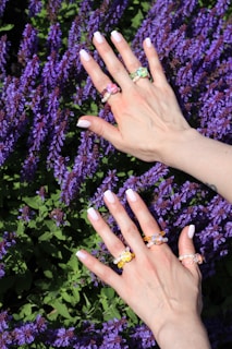 Close-up of hands adorned with pastel nail art and delicate rings resting on a rustic wooden table with wildflowers.