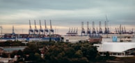 A large industrial port with numerous cranes lining the docks, handling stacks of shipping containers. Wind turbines can be seen on the horizon, indicating a blend of industry and renewable energy. The foreground is dotted with trees and industrial buildings.