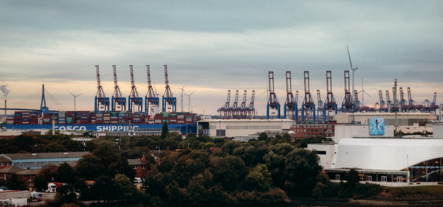 A large industrial port with numerous cranes lining the docks, handling stacks of shipping containers. Wind turbines can be seen on the horizon, indicating a blend of industry and renewable energy. The foreground is dotted with trees and industrial buildings.