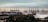 A large industrial port with numerous cranes lining the docks, handling stacks of shipping containers. Wind turbines can be seen on the horizon, indicating a blend of industry and renewable energy. The foreground is dotted with trees and industrial buildings.