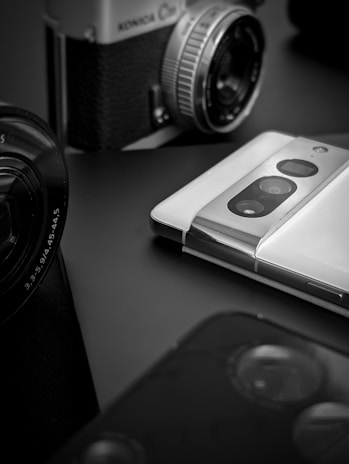 A vintage film camera with a mechanical lens is positioned near a modern smartphone featuring dual rear cameras. The scene is captured in black and white, highlighting the contrast between old and new technology. The focus is primarily on the lens of the camera and the sleek design of the smartphone.