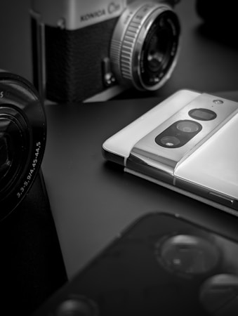 A vintage film camera with a mechanical lens is positioned near a modern smartphone featuring dual rear cameras. The scene is captured in black and white, highlighting the contrast between old and new technology. The focus is primarily on the lens of the camera and the sleek design of the smartphone.