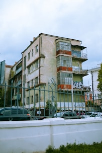An old, worn-out apartment building with peeling paint and graffiti on the walls. The structure has multiple balconies with rusted railings, and some windows are covered with tarps or shutters. Cars are parked in front of the building, and the foreground features an ornate metal fence.