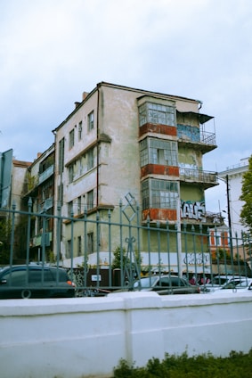 An old, worn-out apartment building with peeling paint and graffiti on the walls. The structure has multiple balconies with rusted railings, and some windows are covered with tarps or shutters. Cars are parked in front of the building, and the foreground features an ornate metal fence.