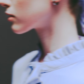 A soft-focus image of a person's lower face and neck, featuring gentle curves and smooth skin. The individual is wearing a light-colored garment with visible folds and ruffles.