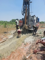 Several individuals are engaged in a work activity involving a large drilling machine set in a rural, grassy area. The setting appears dry, and there is a stream of water or mud being managed by the workers as part of the drilling process. The landscape includes scattered vegetation and a clear blue sky.
