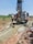 Several individuals are engaged in a work activity involving a large drilling machine set in a rural, grassy area. The setting appears dry, and there is a stream of water or mud being managed by the workers as part of the drilling process. The landscape includes scattered vegetation and a clear blue sky.