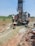 Several individuals are engaged in a work activity involving a large drilling machine set in a rural, grassy area. The setting appears dry, and there is a stream of water or mud being managed by the workers as part of the drilling process. The landscape includes scattered vegetation and a clear blue sky.