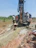 Several individuals are engaged in a work activity involving a large drilling machine set in a rural, grassy area. The setting appears dry, and there is a stream of water or mud being managed by the workers as part of the drilling process. The landscape includes scattered vegetation and a clear blue sky.