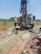 Several individuals are engaged in a work activity involving a large drilling machine set in a rural, grassy area. The setting appears dry, and there is a stream of water or mud being managed by the workers as part of the drilling process. The landscape includes scattered vegetation and a clear blue sky.
