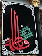 A decorative black banner prominently features ornate Arabic calligraphy in red and green. Intricate white designs frame the banner, adding an elegant touch. The script is set against a dark backdrop, highlighting its colors and details.