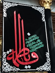 A decorative black banner prominently features ornate Arabic calligraphy in red and green. Intricate white designs frame the banner, adding an elegant touch. The script is set against a dark backdrop, highlighting its colors and details.
