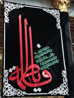 A decorative black banner prominently features ornate Arabic calligraphy in red and green. Intricate white designs frame the banner, adding an elegant touch. The script is set against a dark backdrop, highlighting its colors and details.