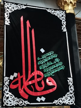 A decorative black banner prominently features ornate Arabic calligraphy in red and green. Intricate white designs frame the banner, adding an elegant touch. The script is set against a dark backdrop, highlighting its colors and details.