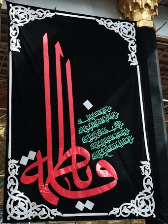 A decorative black banner prominently features ornate Arabic calligraphy in red and green. Intricate white designs frame the banner, adding an elegant touch. The script is set against a dark backdrop, highlighting its colors and details.