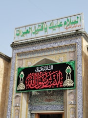 A decorative entrance to a building adorned with intricate blue and gold tiles, Arabic calligraphy, and an ornate sign with red and green script. A large sign in Arabic is displayed prominently above the door, surrounded by geometric and floral patterns.