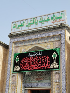 A decorative entrance to a building adorned with intricate blue and gold tiles, Arabic calligraphy, and an ornate sign with red and green script. A large sign in Arabic is displayed prominently above the door, surrounded by geometric and floral patterns.