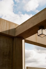 Concrete architectural structures intersect with each other, creating a geometric design against a backdrop of a partly cloudy sky. A cubic light fixture is suspended from one of the beams.