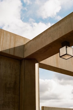 Concrete architectural structures intersect with each other, creating a geometric design against a backdrop of a partly cloudy sky. A cubic light fixture is suspended from one of the beams.