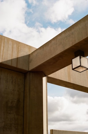 Concrete architectural structures intersect with each other, creating a geometric design against a backdrop of a partly cloudy sky. A cubic light fixture is suspended from one of the beams.